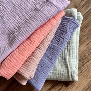 A stack of muslin fabric baby blankets in pastel colors—lavender, coral, dusty pink, denim blue, and sage green